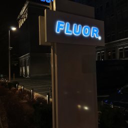 Auror Stacja Kontroli Reklamy Marek Krzywda - Iluminowany szyld z nazwą firmy Fluor na tle budynku biurowego nocą, z widocznymi elementami parkingu i oświetlenia zewnętrznego.