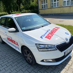 Auror Stacja Kontroli Reklamy Marek Krzywda - Biały samochód z logo pizzerii 'Bombola' i numerem telefonu, zaparkowany na chodniku z kostki brukowej przed budynkiem.