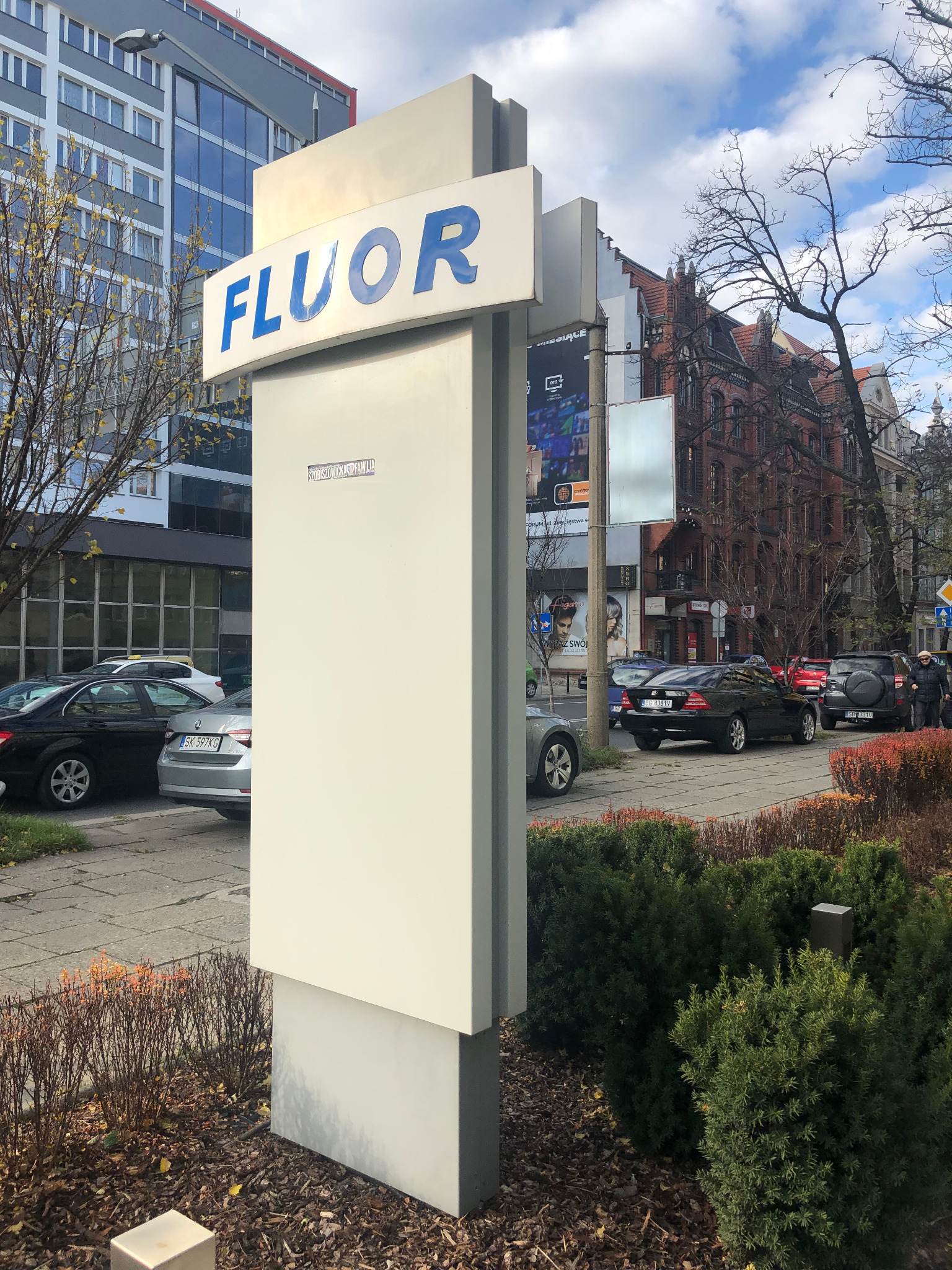 Pylon reklamowy z napisem 'FLUOR' w niebieskim kolorze, umieszczony na tle miejskiego krajobrazu z budynkami i samochodami.