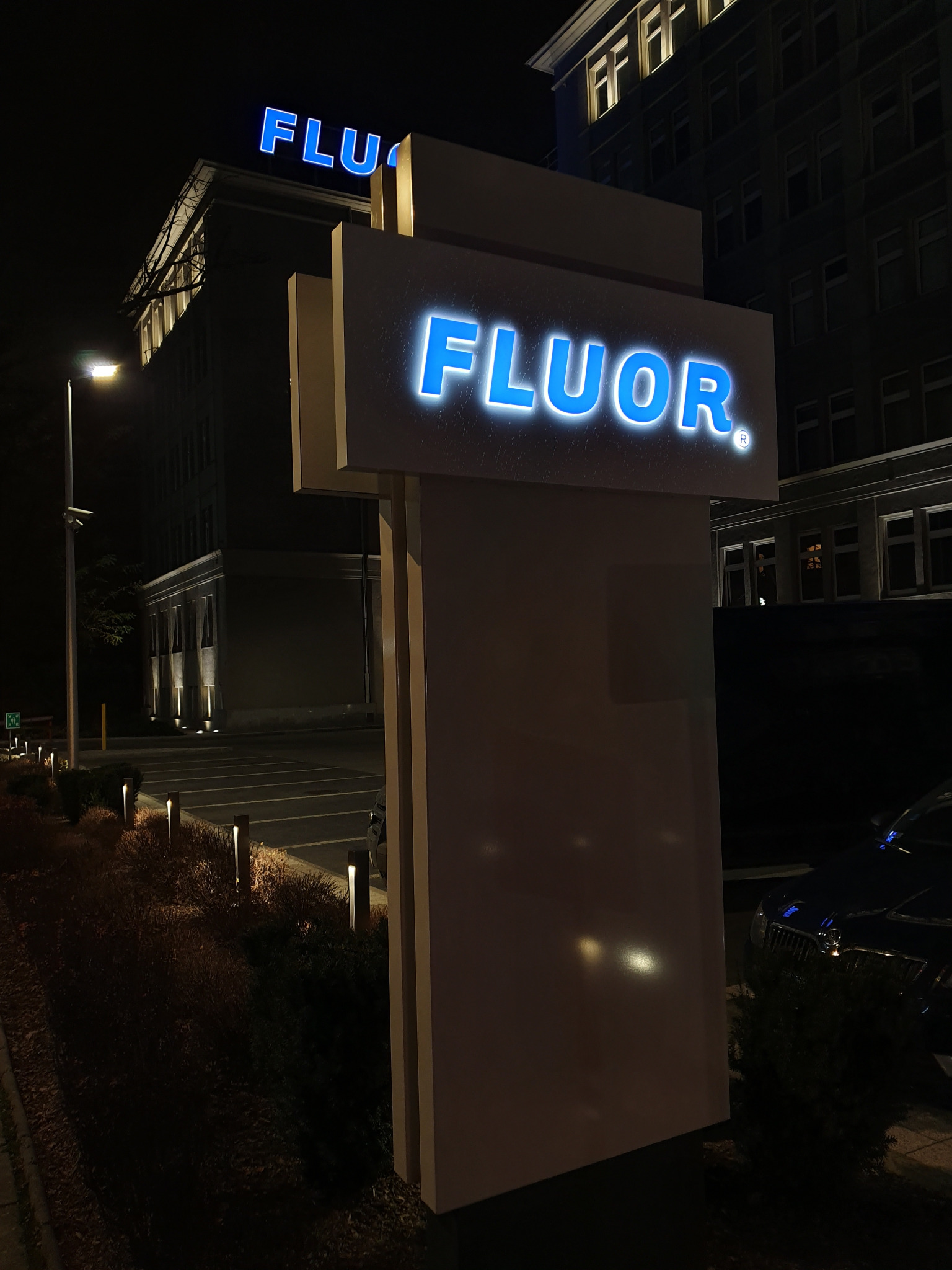 Iluminowany szyld z nazwą firmy Fluor na tle budynku biurowego nocą, z widocznymi elementami parkingu i oświetlenia zewnętrznego.