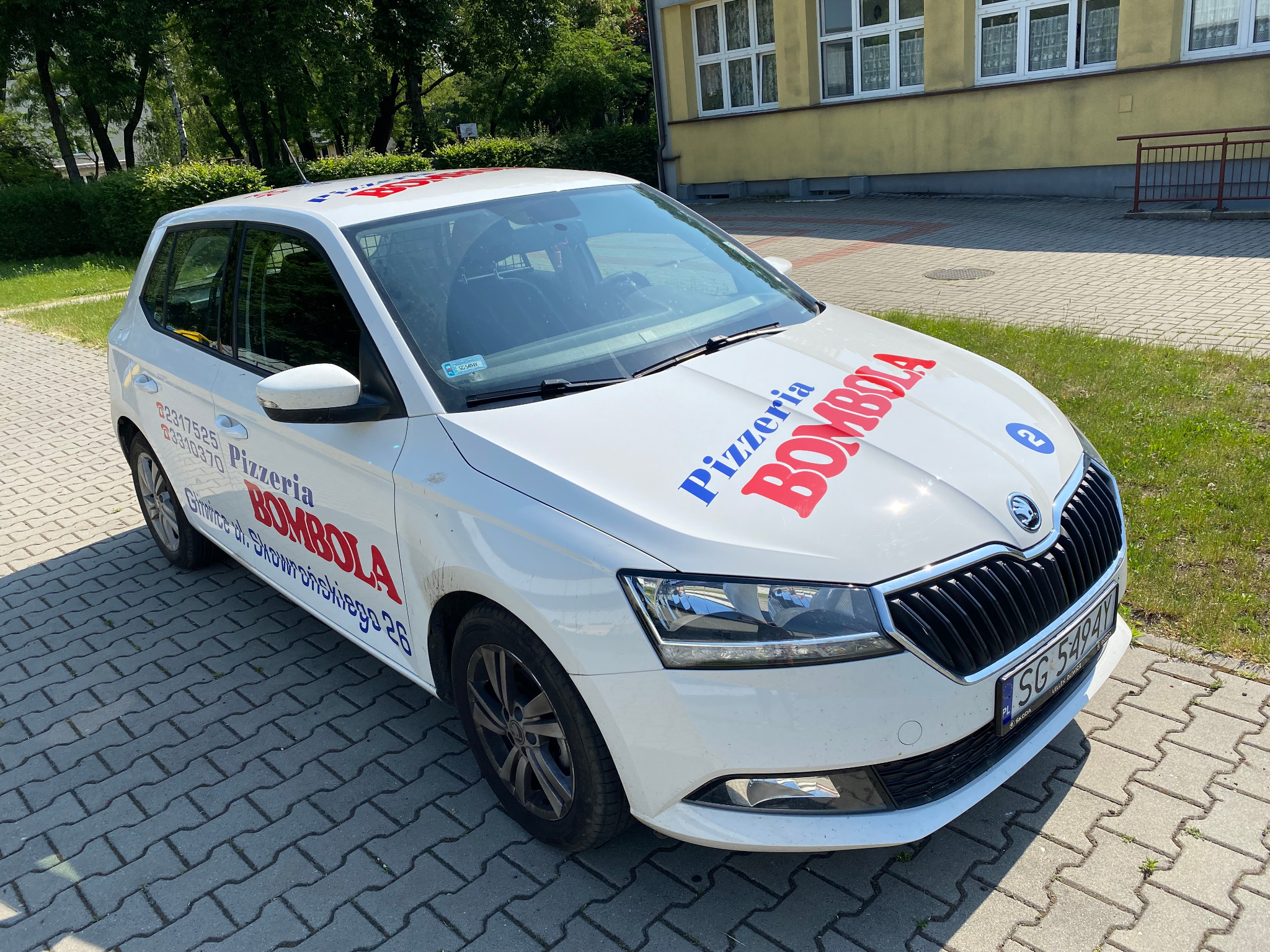 Biały samochód z logo pizzerii 'Bombola' i numerem telefonu, zaparkowany na chodniku z kostki brukowej przed budynkiem.