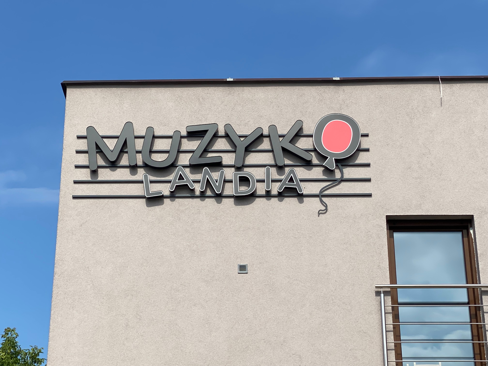 Zewnętrzny szyld reklamowy z nazwą 'Muzykolandia' i motywem balonu umieszczony na elewacji budynku z oknem i balkonem, błękitne niebo w tle.