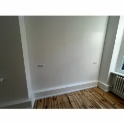 Wnętrze pokoju po odświeżeniu ścian, widoczne gniazdka elektryczne, drewniana podłoga i białe listwy przypodłogowe oraz grzejnik pod oknem.