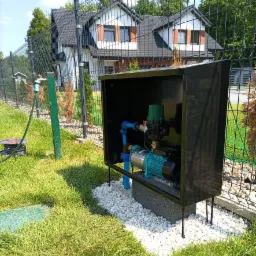 Zewnętrzna instalacja pompy do wody w czarnej obudowie, otoczonej białym żwirem, na tle trawnika i ogrodzenia z widocznym domem w oddali.