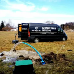 Test wydajności studni: woda tryska z niebieskiej rury podłączonej do studni z zieloną pokrywą na tle czarnego samochodu dostawczego z napisem 'WIERCENIE STUDNI' i sprzętu do wiercenia.