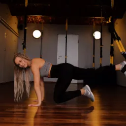 Kobieta w sportowym stroju ćwiczy z taśmami TRX w siłowni, widoczne drewniane podłogi i ceglany sufit.