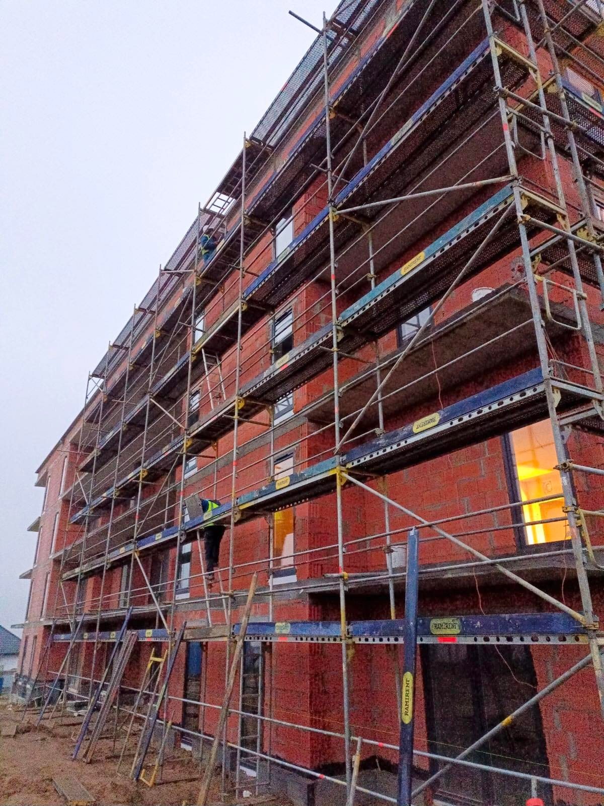 Budynek w trakcie budowy, z widocznymi rusztowaniami i pracownikiem w kamizelce odblaskowej na jednym z poziomów, ceglana elewacja w surowym stanie, pochmurne niebo w tle.