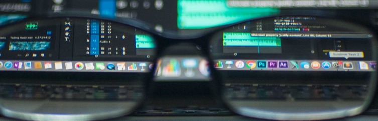 Ekran laptopa z widocznym interfejsem edytora audio i kodu, widziany przez szkła okularów, z widocznymi ikonami Adobe Photoshop, Premiere i After Effects.