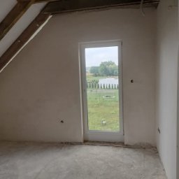 Damian Łojek - Usługi Tynkarskie - Poddasze w surowym stanie po nałożeniu tynku, widoczne drewniane elementy konstrukcji dachu, okno z widokiem na jezioro i trawnik, ściany przygotowane do dalszych prac wykończeniowych.