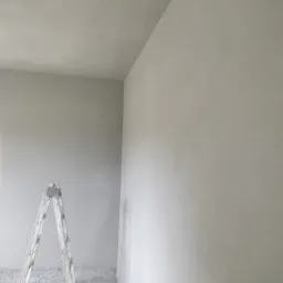 Świeżo otynkowane wnętrze z aluminiową drabiną i wiadrem na podłodze, widoczne niedoskonałości wykończenia przy podłodze.