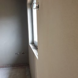 Damian Łojek - Usługi Tynkarskie - Świeżo otynkowana ściana w szaro-beżowym odcieniu, widoczne gniazdka elektryczne i okno z parapetem, surowa podłoga, brak wykończenia.