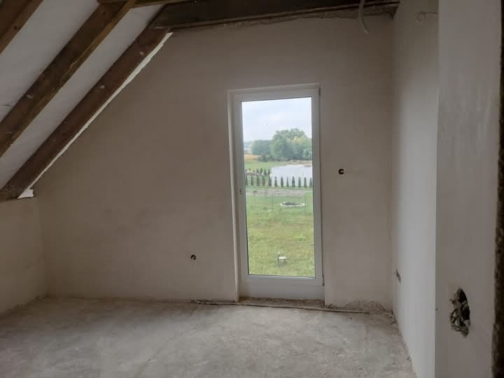 Poddasze w surowym stanie po nałożeniu tynku, widoczne drewniane elementy konstrukcji dachu, okno z widokiem na jezioro i trawnik, ściany przygotowane do dalszych prac wykończeniowych.