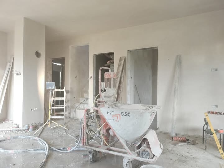 Wnętrze w trakcie prac wykończeniowych: ściany pokryte świeżym tynkiem, widoczna maszyna do tynkowania z wyraźnym napisem 'PFT G5c', drabina z oświetleniem halogenowym oraz poziomica oparta o krzesło.