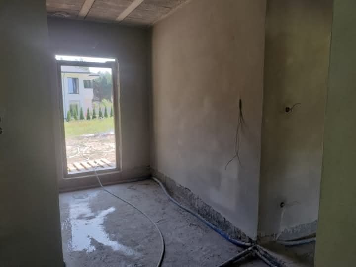 Wnętrze pomieszczenia w trakcie prac wykończeniowych, ściany pokryte świeżym tynkiem, widoczne wystające kable elektryczne, na podłodze resztki zaprawy i węże doprowadzające wodę.