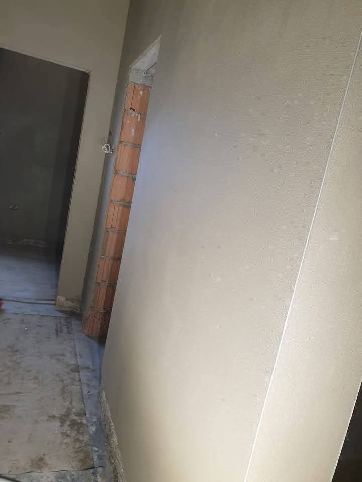 Świeżo otynkowana ściana wewnątrz budynku, z widocznym fragmentem muru z czerwonej cegły przy otworze drzwiowym i szarą podłogą w tle.