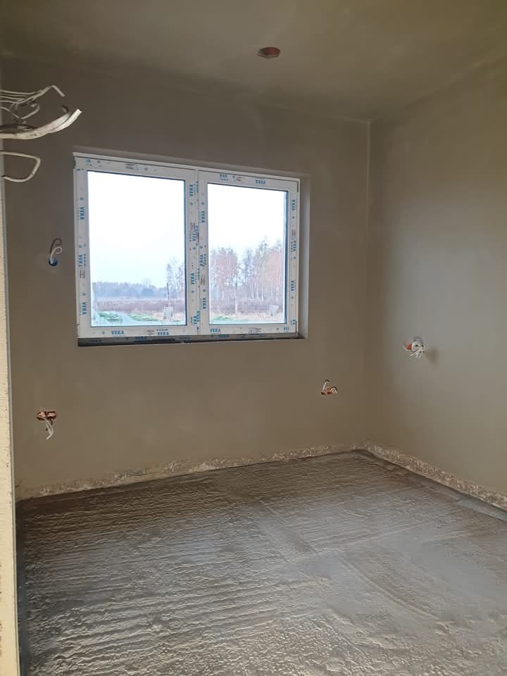 Świeżo wykonane, surowe wykończenie ścian i posadzki w pomieszczeniu z białym oknem PCV, widoczne wystające kable elektryczne.