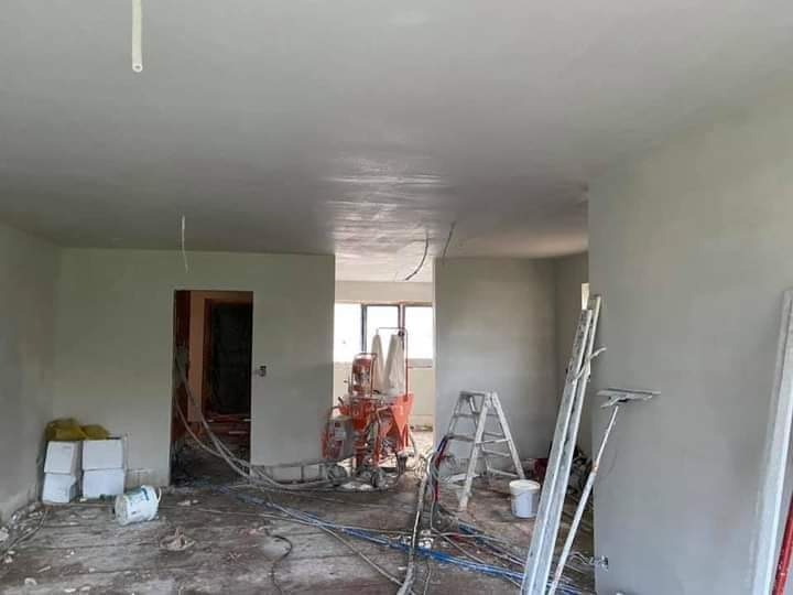 Wnętrze w trakcie prac wykończeniowych: świeżo położone tynki na ścianach i suficie, widoczna maszyna do tynkowania, drabina i narzędzia malarskie na podłodze pokrytej kablami.