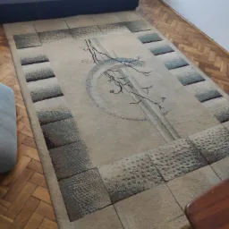 Beżowy dywan z geometrycznym wzorem i motywem gałązek na parkiecie, fragment sofy w tle.