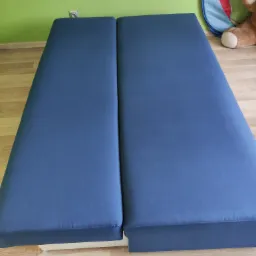 Dwa połączone materace w niebieskim pokrowcu na drewnianej podłodze, widoczny fragment ściany w kolorze limonkowym z gniazdkiem elektrycznym oraz zabawki w tle.