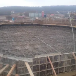 Przygotowanie zbrojenia fundamentu o nietypowym kształcie w Katowicach, widoczne szalunki i siatka zbrojeniowa, pracownik w kasku i kamizelce odblaskowej kontroluje postęp prac.