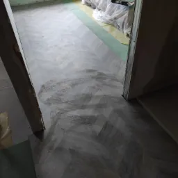 Ułożona podłoga z paneli w wzór jodełki francuskiej, widoczne paczki paneli w folii ochronnej w tle, częściowo widoczne podłoże pod panele.