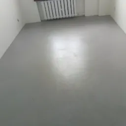 Szara, gładka podłoga w pustym pokoju z białymi ścianami i grzejnikiem pod oknem.