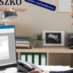 Kobieta przy biurku z komputerem, na ekranie widoczny program księgowy, w tle biurowe akcesoria i dekoracje, nad biurkiem logo firmy.