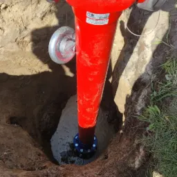 Jaskrawoczerwony hydrant przeciwpożarowy, częściowo zagłębiony w ziemi, z widoczną niebieską podstawą i świeżo zasypanym wykopem, otoczony trawą.