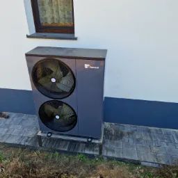 Szara pompa ciepła Kensol z dwoma wentylatorami, zamontowana na metalowej podstawie przy ścianie budynku z widocznym oknem.