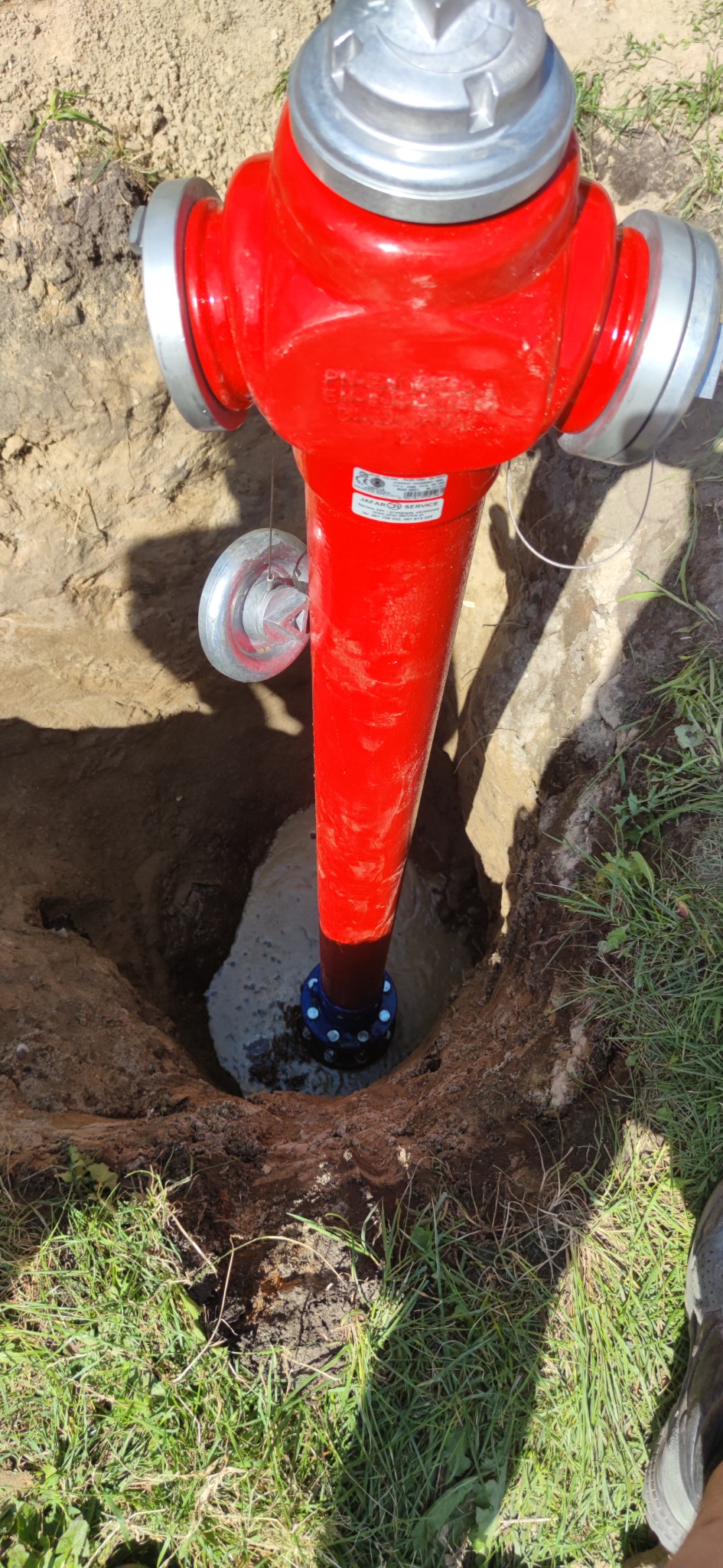Jaskrawoczerwony hydrant przeciwpożarowy, częściowo zagłębiony w ziemi, z widoczną niebieską podstawą i świeżo zasypanym wykopem, otoczony trawą.