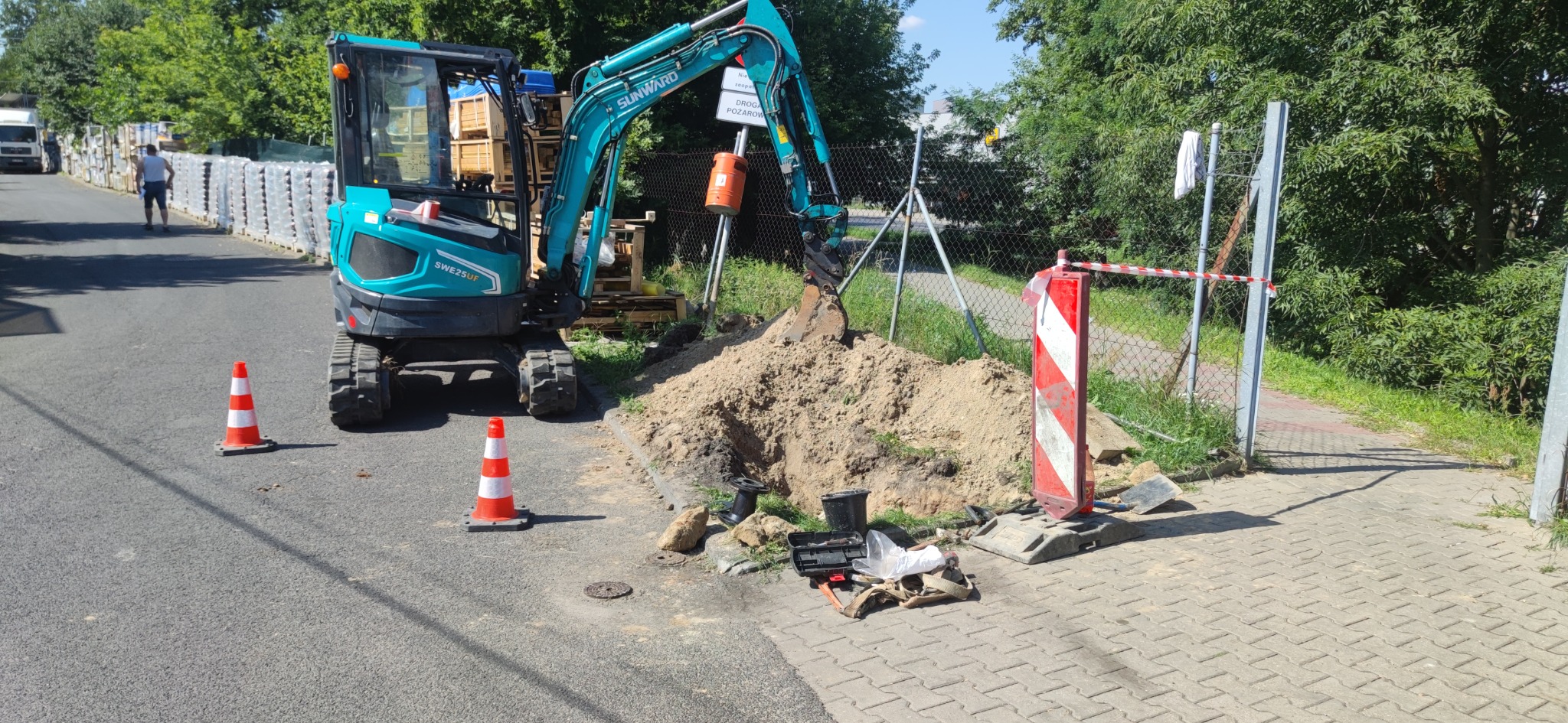 Niebieska mini koparka Sunward SWE25UF wykonuje wykop w ziemi przy ulicy, zabezpieczony znakami ostrzegawczymi i pachołkami, widoczne narzędzia i akcesoria hydrauliczne.