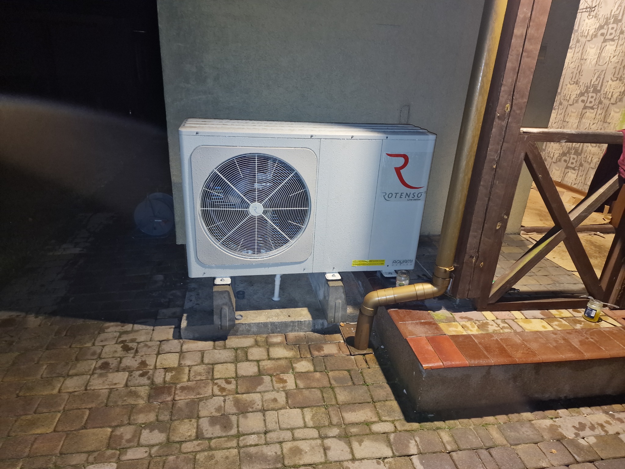 Zewnętrzna jednostka pompy ciepła Rotenso Aquami Series, zamontowana na betonowych podstawach na brukowanym patio, widoczna rura odprowadzająca skropliny, ujęcie nocne.
