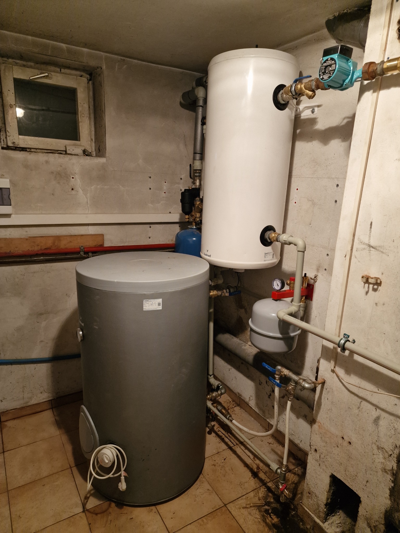 Dwa zbiorniki na wodę (biały i szary) w piwnicy z widocznymi rurami, zaworami, manometrem i pompą obiegową. Ściany i podłoga wykończone w surowym stylu.