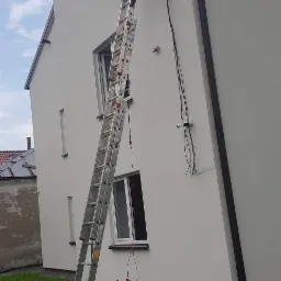 Pracownik na drabinie, wiercący otwór w elewacji budynku pod montaż anteny satelitarnej, widoczna antena na trawie, skrzynka z narzędziami przy drabinie.