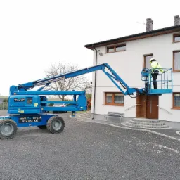 Niebieski wózek widłowy teleskopowy z operatorem w żółtej kamizelce, podnośnik koszowy przy białym budynku z drewnianymi oknami, szare podłoże z kamyków.