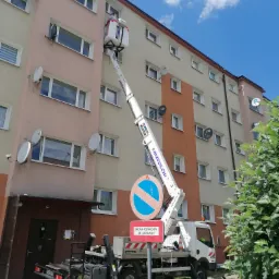 Podnośnik koszowy marki Bizzocchi na małym samochodzie ciężarowym, ustawiony przed blokiem mieszkalnym z antenami satelitarnymi, znak zakazu zatrzymywania i postoju z informacją o drodze pożarowej.