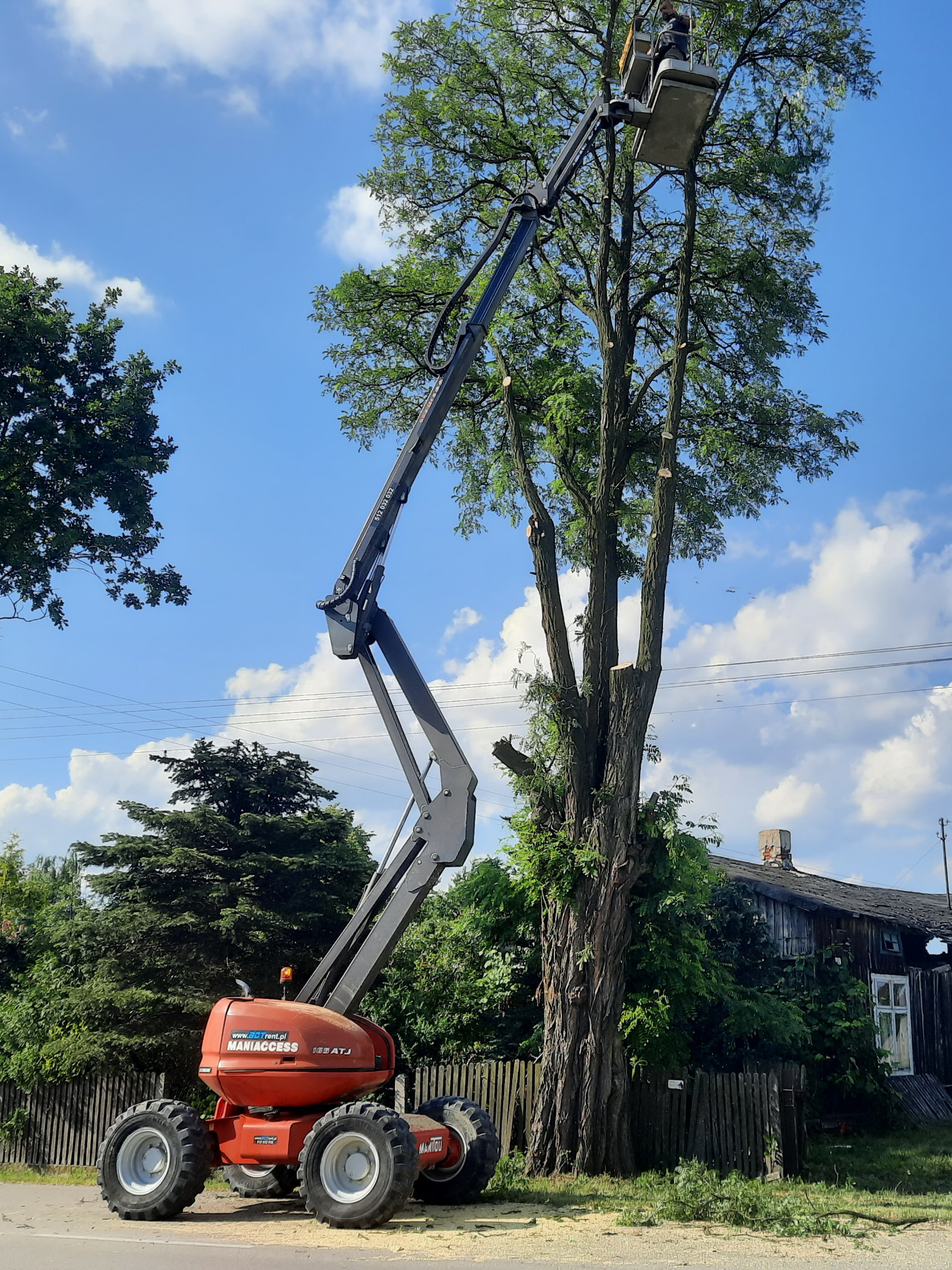 Pracownik w koszu podnośnika Maniaccess 163 ATJ, przycina gałęzie wysokiego drzewa w pobliżu drewnianego domu z uszkodzonym dachem, widoczne ślady po cięciu gałęzi.