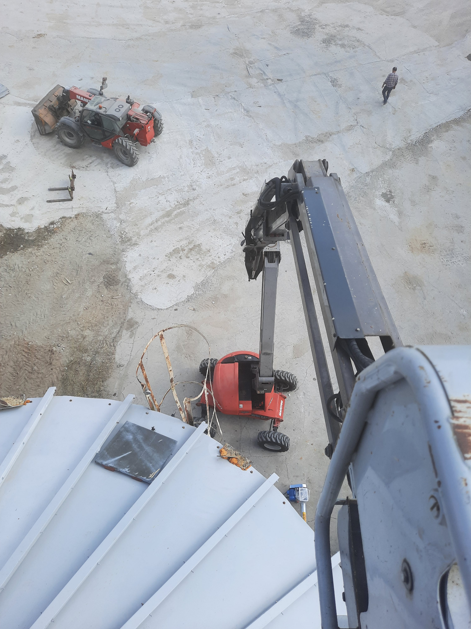 Widok z góry na plac budowy z dwoma wózkami widłowymi Manitou, jednym z łyżką, drugim z osprzętem roboczym, oraz pojedynczą osobą w oddali, z perspektywy podnośnika koszowego nad konstrukcją dachu.