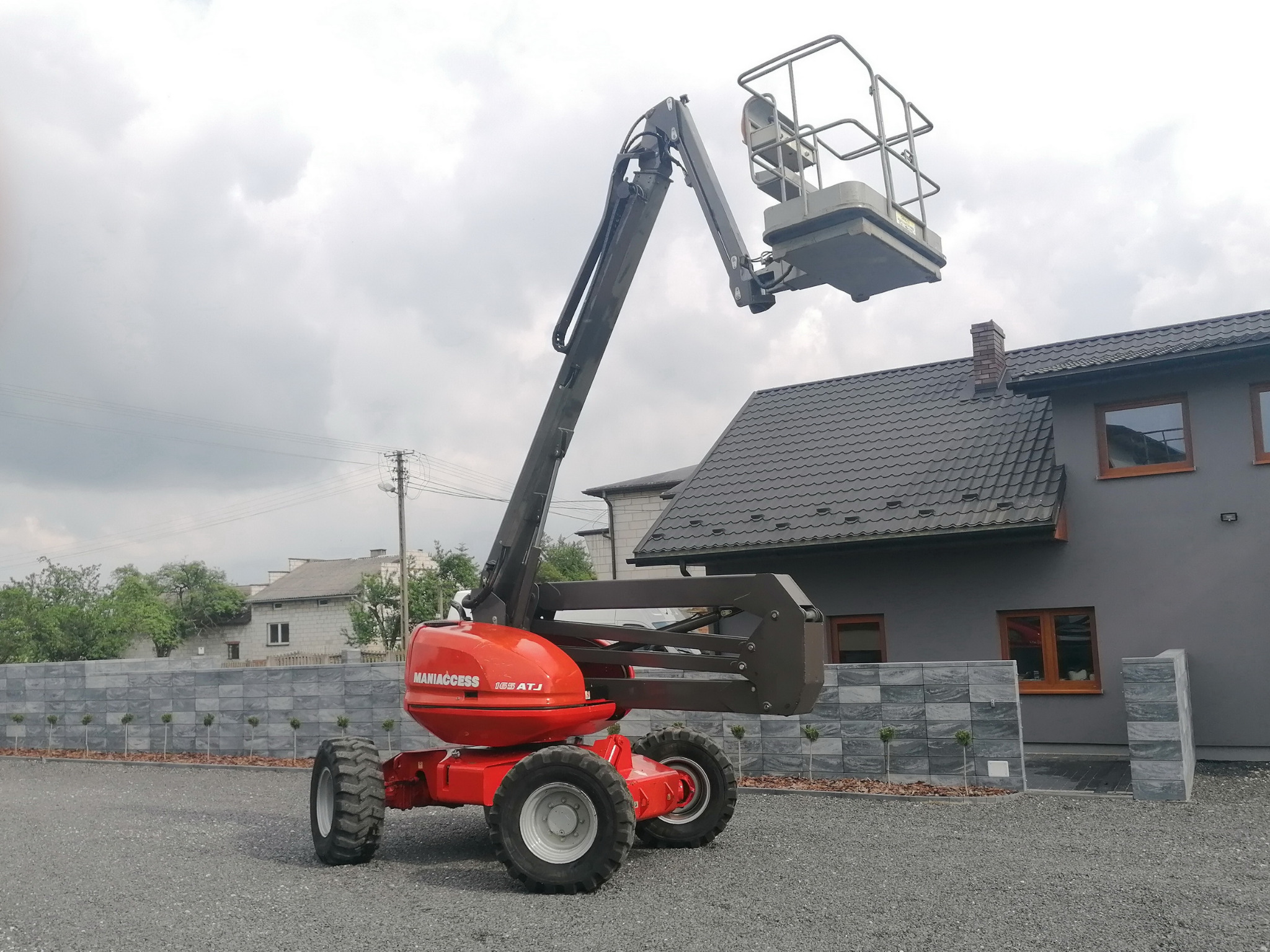 Czerwony wózek widłowy Manitou 165 ATJ na żwirowym podłożu, z podniesionym koszem na tle szarego domu i pochmurnego nieba.