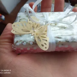 Ręcznie robione mydło zapakowane w folię z różowymi kropkami, przewiązane białą wstążką i ozdobione papierowym motylem z perłą w środku, prezentowane na dłoni.