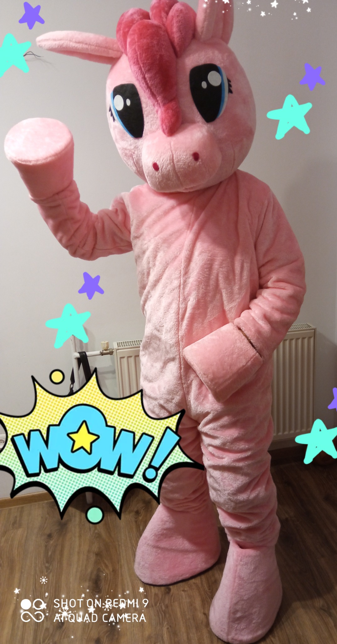 Różowy kostium maskotki jednorożca z dużymi oczami, stojący na drewnianej podłodze z dodanymi cyfrowo gwiazdkami i napisem 'WOW!'