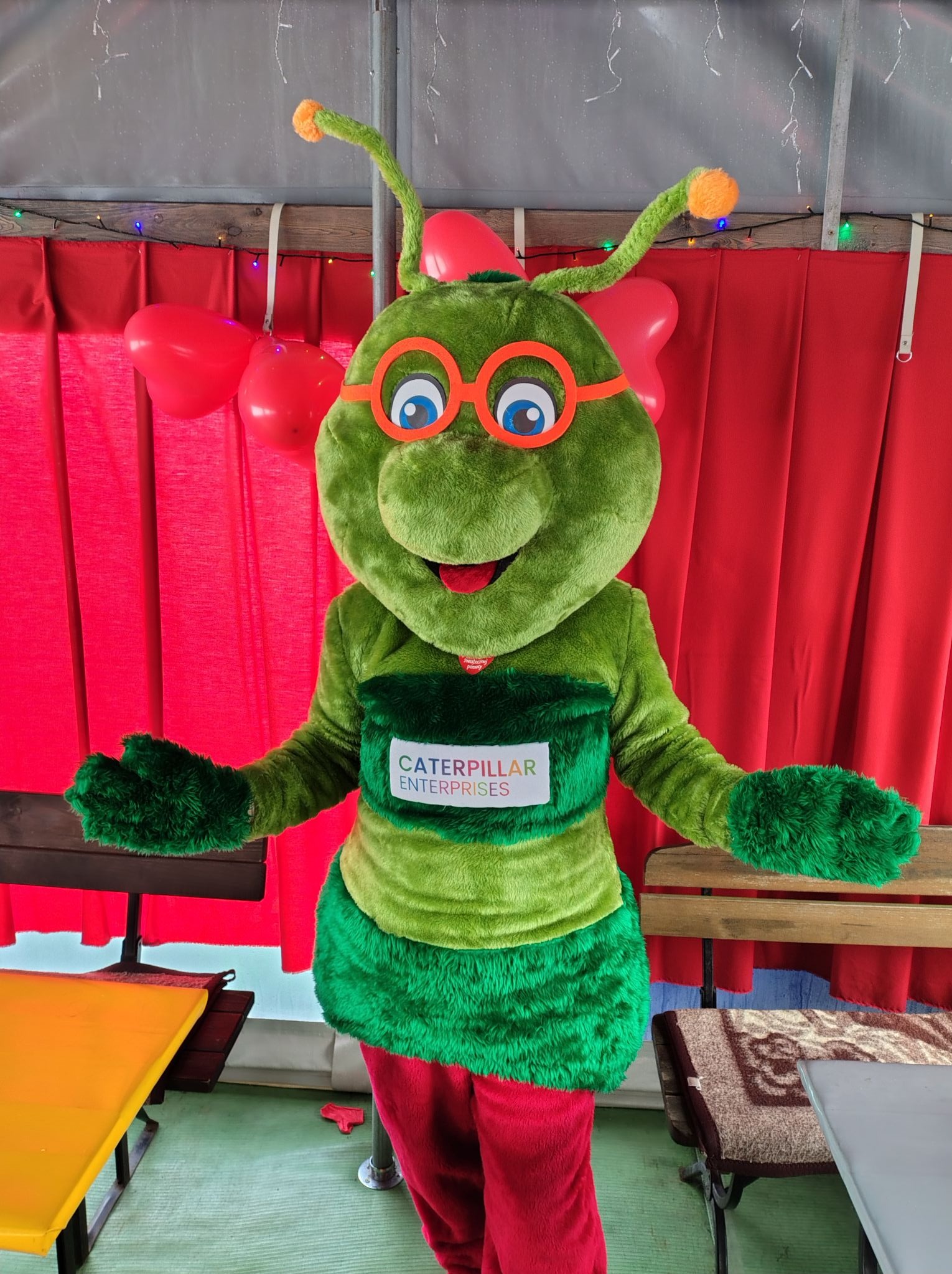 Uśmiechnięty, pluszowy kostium zielonej gąsienicy z pomarańczowymi okularami i czułkami, prezentujący się na tle czerwonych dekoracji w kształcie serc i firmowego logo 'Caterpillar Enterprises'.
