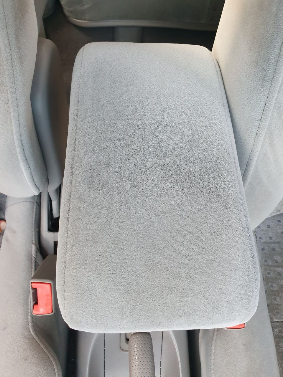 Szary, welurowy podłokietnik samochodowy widziany z góry, z fragmentem fotela i pasem bezpieczeństwa.