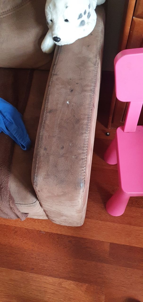 Brązowy, zabrudzony podłokietnik sofy z pluszowym psem na górze i różowym krzesełkiem dziecięcym obok, na tle drewnianej podłogi.