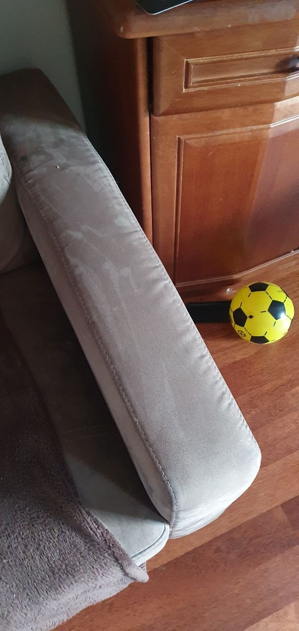 Fragment beżowego, pluszowego fotela z widocznym szwem na podłokietniku, obok żółto-czarna piłka do gry i fragment drewnianej komody.