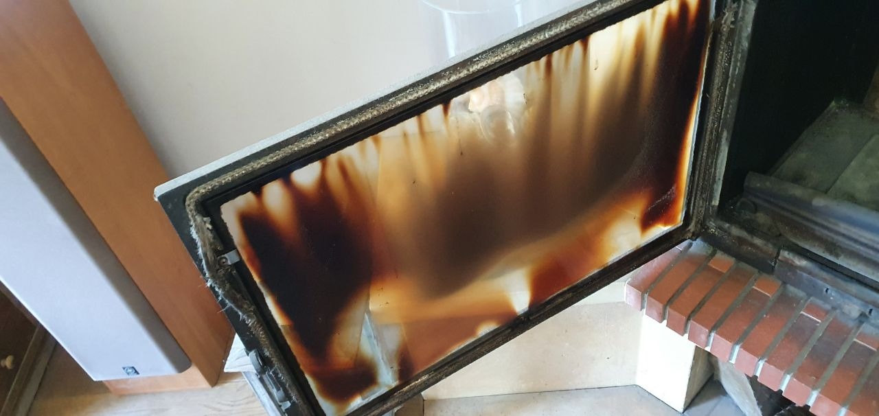 Brudna szyba kominkowa z widocznymi smugami sadzy i osadu po spalaniu, uchylona na tle ceglanej obudowy kominka.