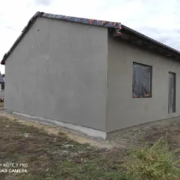 Świeżo otynkowany, parterowy dom jednorodzinny z trzema oknami zabezpieczonymi folią, widok z boku na tle pochmurnego nieba. W tle sterta drewna i sąsiedni dom.