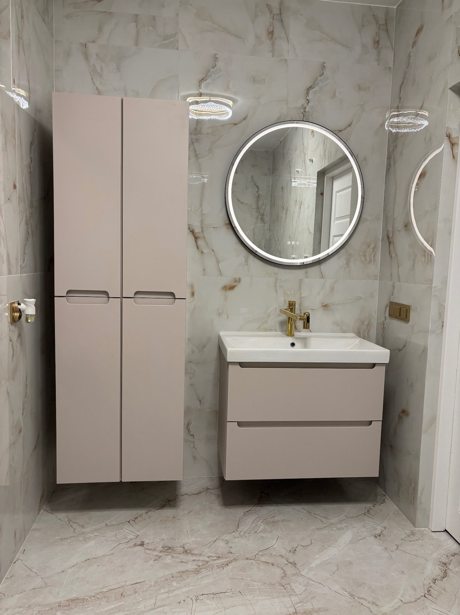 Jasna łazienka z marmurowymi płytkami, beżowymi szafkami i okrągłym lustrem z podświetleniem LED. Złota bateria umywalkowa dodaje elegancji.