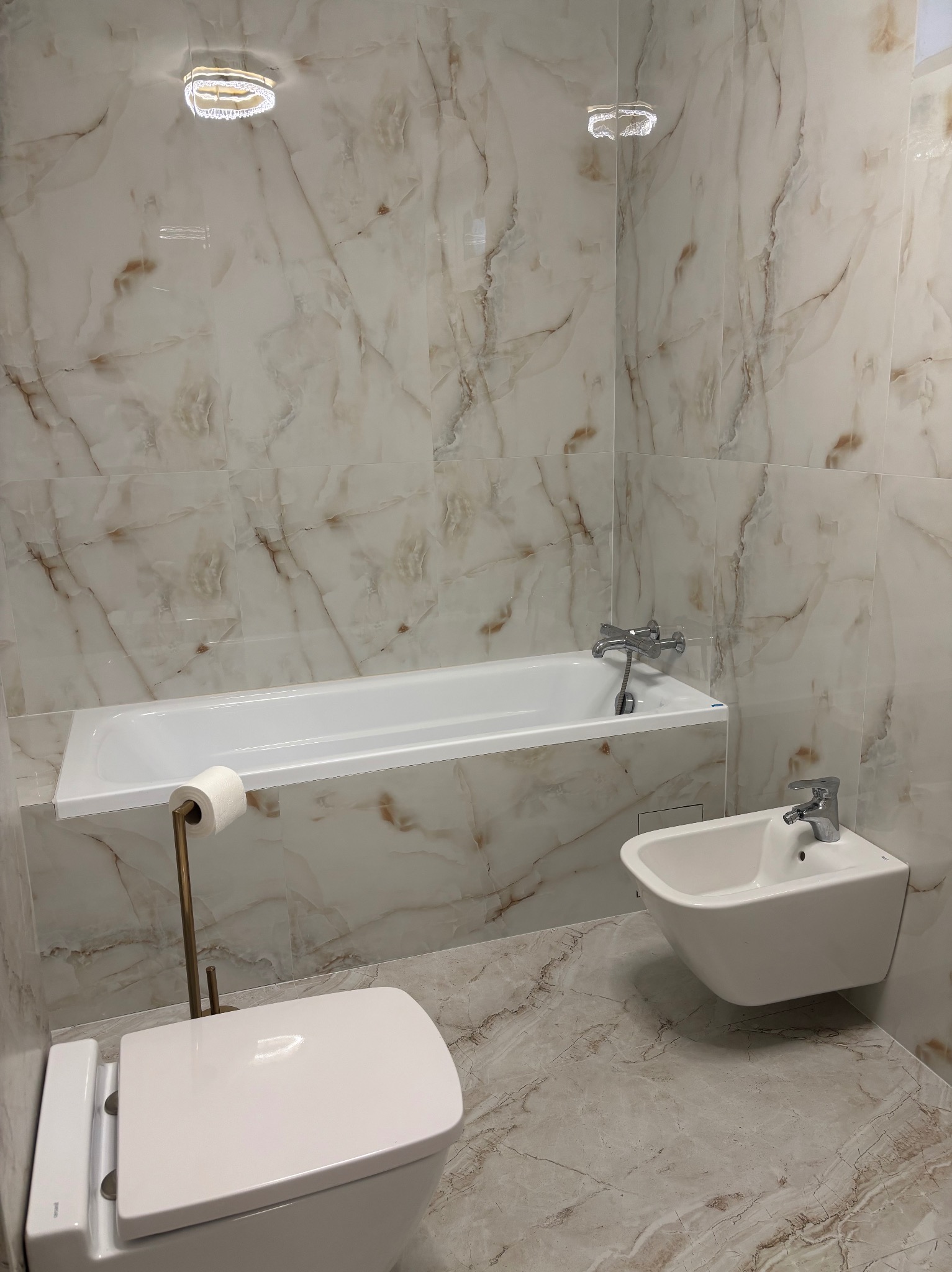 Elegancka łazienka w beżowych marmurach: wanna, bidet i toaleta. Oświetlenie sufitowe podkreśla luksusowy charakter wykończenia. Widok na całe pomieszczenie.