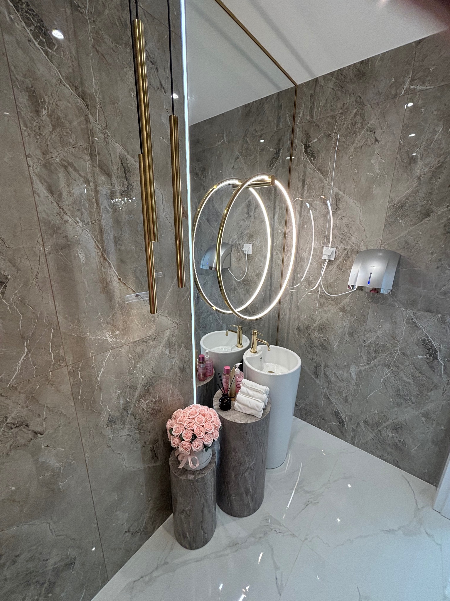 Elegancka łazienka z marmurowymi płytkami, podwójną umywalką na postumentach, okrągłym lustrem z podświetleniem LED i złotymi lampami wiszącymi.